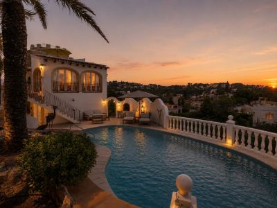 Exterior de la villa con piscina curva y balustrada iluminada, junto a una zona de estar al aire libre y ventanas arqueadas, todo bajo el cálido resplandor del atardecer.