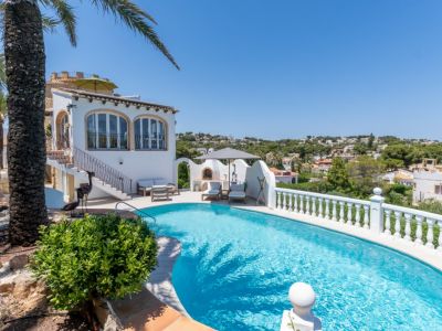 La piscina curva de agua azul cristalina se integra con la villa mediterránea blanca, que presenta ventanas arqueadas y una zona de barbacoa exterior con tumbonas, en un entorno verde.