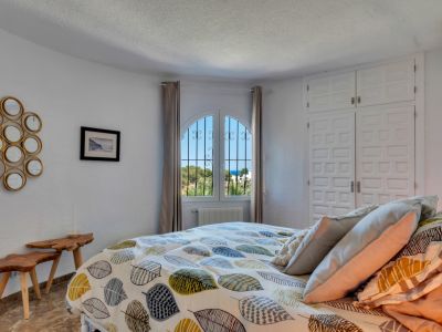 Luminoso dormitorio con ventana arqueada y rejas de hierro forjado, que ofrece vistas parciales al mar. Cuenta con armarios empotrados blancos y mesas auxiliares de madera rústica.