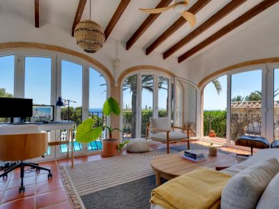 Luminoso salón con suelo de terracota y vigas de madera, que ofrece un espacio de trabajo con vistas panorámicas al mar y la piscina a través de sus ventanales arqueados.