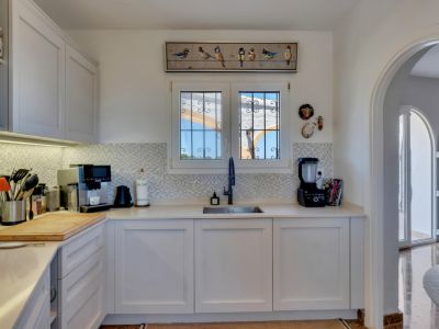 Moderna cocina con armarios blancos, encimeras claras y salpicadero de mosaico. Una ventana con rejas ofrece vistas al exterior, y un arco conduce a un espacio adyacente luminoso.