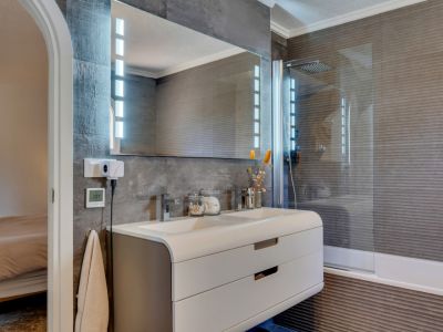 Moderno baño con doble lavabo y mueble curvo, espejo iluminado con LED y paredes de azulejos texturizados en gris oscuro. La ducha integrada con mampara de cristal complementa el diseño.