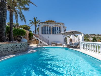 Piscina curva de agua azul con zona de solárium, barbacoa exterior y una villa mediterránea de fachada blanca, rodeada de palmeras y vegetación.