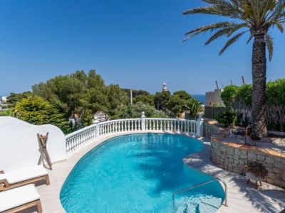 Piscina de diseño curvo con balaustrada blanca, tumbonas y palmera. Ofrece vistas lejanas al mar y un faro bajo un cielo despejado.