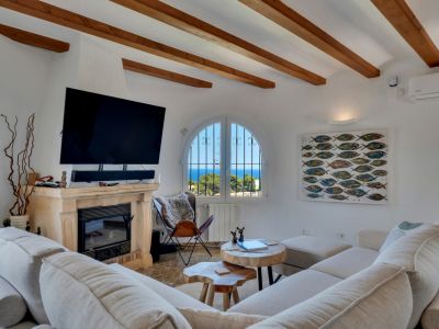 Salón con vigas de madera a la vista y chimenea de piedra, ofreciendo vistas al mar a través de una ventana arqueada. Sofá modular y mesas de centro de madera natural.