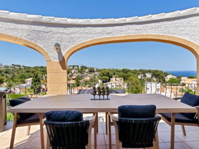 Terraza cubierta con mesa de comedor de madera clara y sillas con cojines oscuros, ofreciendo vistas panorámicas al mar y al paisaje urbano a través de arcos mediterráneos.