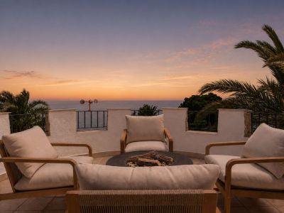Terraza en la azotea con cuatro sillas de madera y cojines claros alrededor de una hoguera, ofreciendo vistas panorámicas al mar y palmeras bajo un cielo de atardecer vibrante.