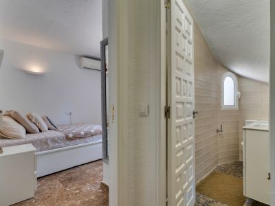 Vista a un dormitorio con suelo efecto mármol y aire acondicionado, frente a un baño con paredes alicatadas en beige, ventana arqueada y suelo de mosaico con lavabo blanco.