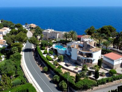 Vista aérea de la villa mediterránea en Jávea, con piscina elevada, jardines aterrazados y arquitectura tradicional, frente al vasto mar azul y un faro lejano.