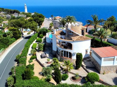 Vista aérea de una villa mediterránea con paredes blancas, tejado de tejas y piscina. Jardines exuberantes rodean la propiedad, con el mar y un faro al fondo.