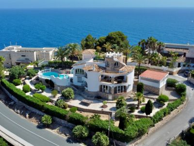 Vista aérea de una villa mediterránea con torre circular, jardines escalonados y piscina de forma libre, todo con el mar azul de fondo.
