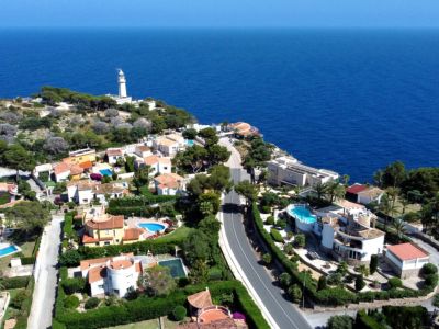 Vista aérea panorámica de una urbanización costera con villas, piscinas privadas y jardines exuberantes, bordeando el azul profundo del Mediterráneo y un faro lejano.