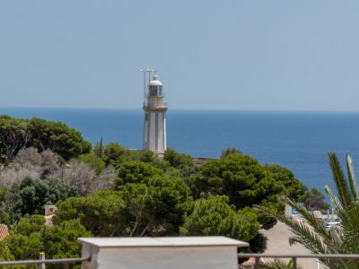 Vista del faro blanco con rayas grises sobre una colina verde, enmarcado por el Mediterráneo azul y un cielo despejado, con vegetación exuberante en primer plano.