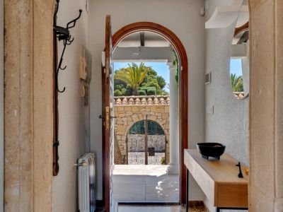 Vista desde el recibidor a través de una puerta de madera arqueada hacia un muro de piedra con verja de hierro forjado y exuberante vegetación mediterránea. En el interior, consola de madera y espejo.