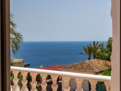 Vista desde un balcón con balaustrada blanca, tejado de terracota y palmeras, todo con vistas al vasto mar Mediterráneo y un velero en la distancia.
