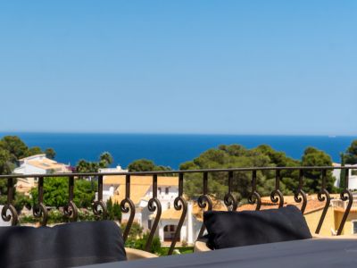 Vista desde una terraza con barandilla de hierro forjado, mostrando tejados de terracota entre pinos y el mar Mediterráneo.