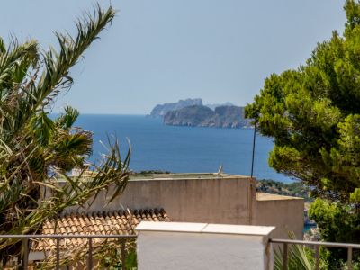 Vista panorámica al mar con costa rocosa distante, enmarcada por palmeras y pinos. Tejas de terracota tradicionales son visibles abajo.