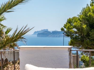Vista panorámica al mar con una isla montañosa en el horizonte, enmarcada por vegetación mediterránea y una barandilla de terraza.