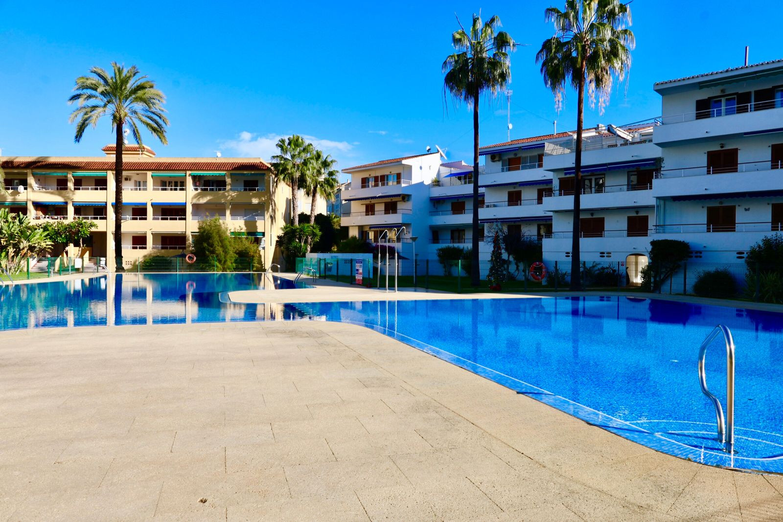Amplia piscina comunitaria con azulejos azules, rodeada de un solárium de baldosas claras y palmeras. Edificios residenciales con balcones y toldos azules al fondo bajo un cielo despejado. Amplia piscina comunitaria con azulejos azules, rodeada de un solárium de baldosas claras y palmeras. Edificios residenciales con balcones y toldos azules al fondo bajo un cielo despejado.
