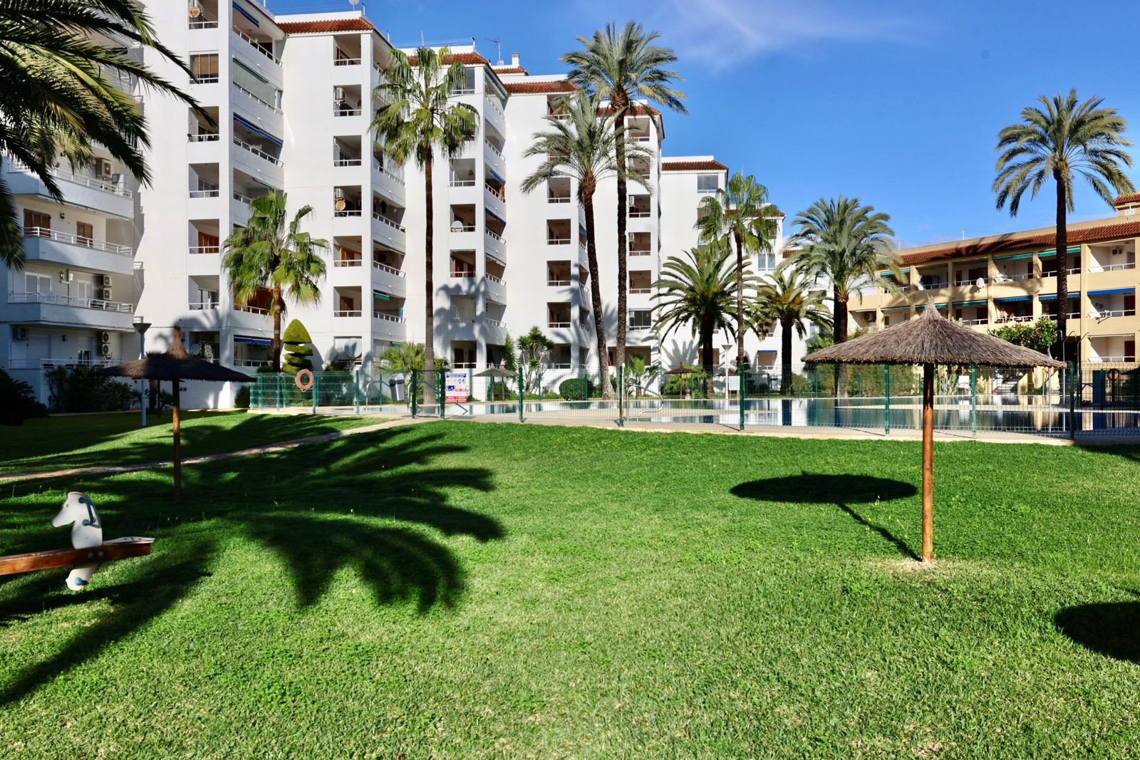 Amplia zona ajardinada comunitaria con césped verde, palmeras y sombrillas de paja. Al fondo, edificios residenciales con balcones y una piscina vallada. Amplia zona ajardinada comunitaria con césped verde, palmeras y sombrillas de paja. Al fondo, edificios residenciales con balcones y una piscina vallada.