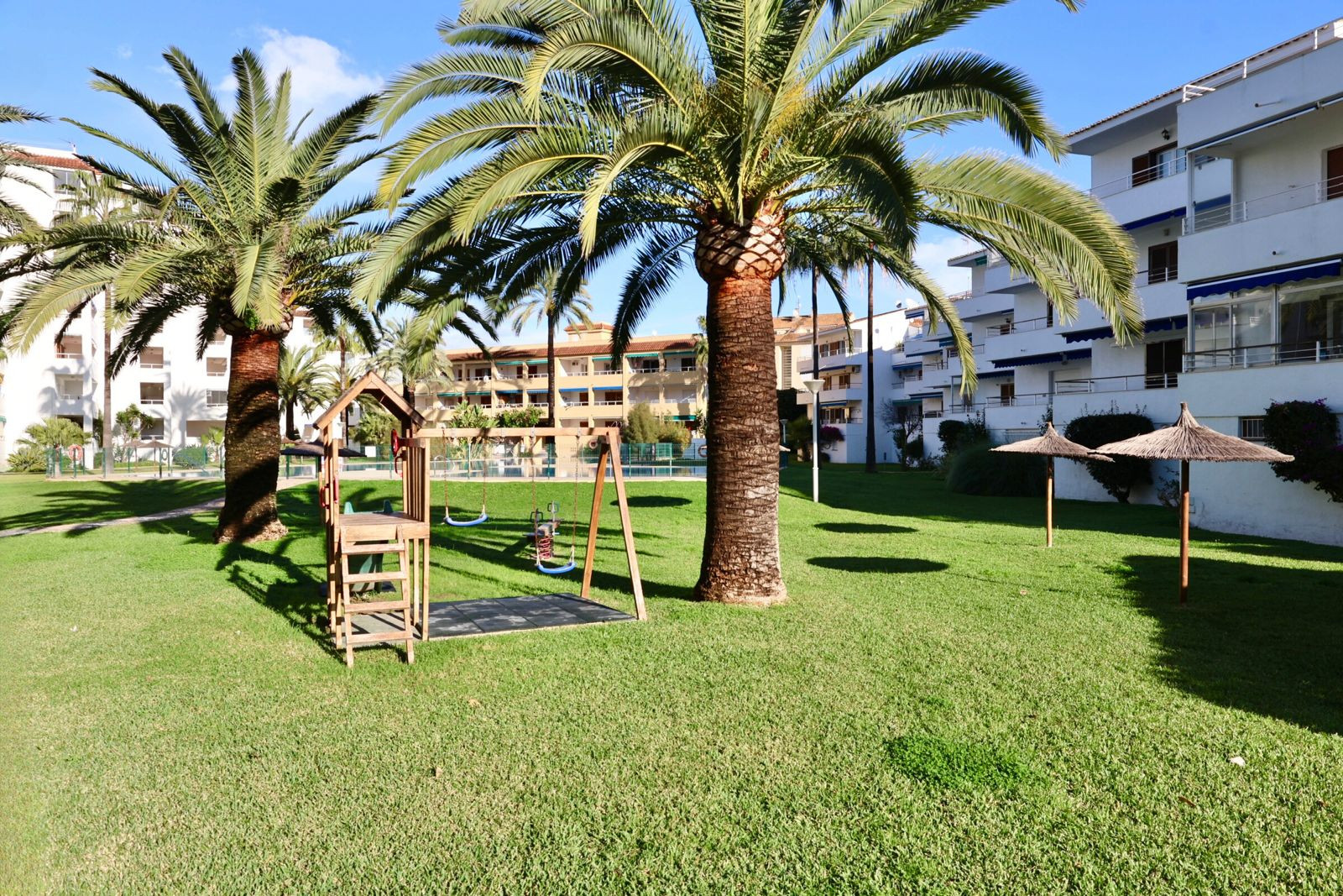 Amplio jardín comunitario con césped cuidado, parque infantil de madera, palmeras y sombrillas de paja, rodeado de edificios residenciales. Amplio jardín comunitario con césped cuidado, parque infantil de madera, palmeras y sombrillas de paja, rodeado de edificios residenciales.