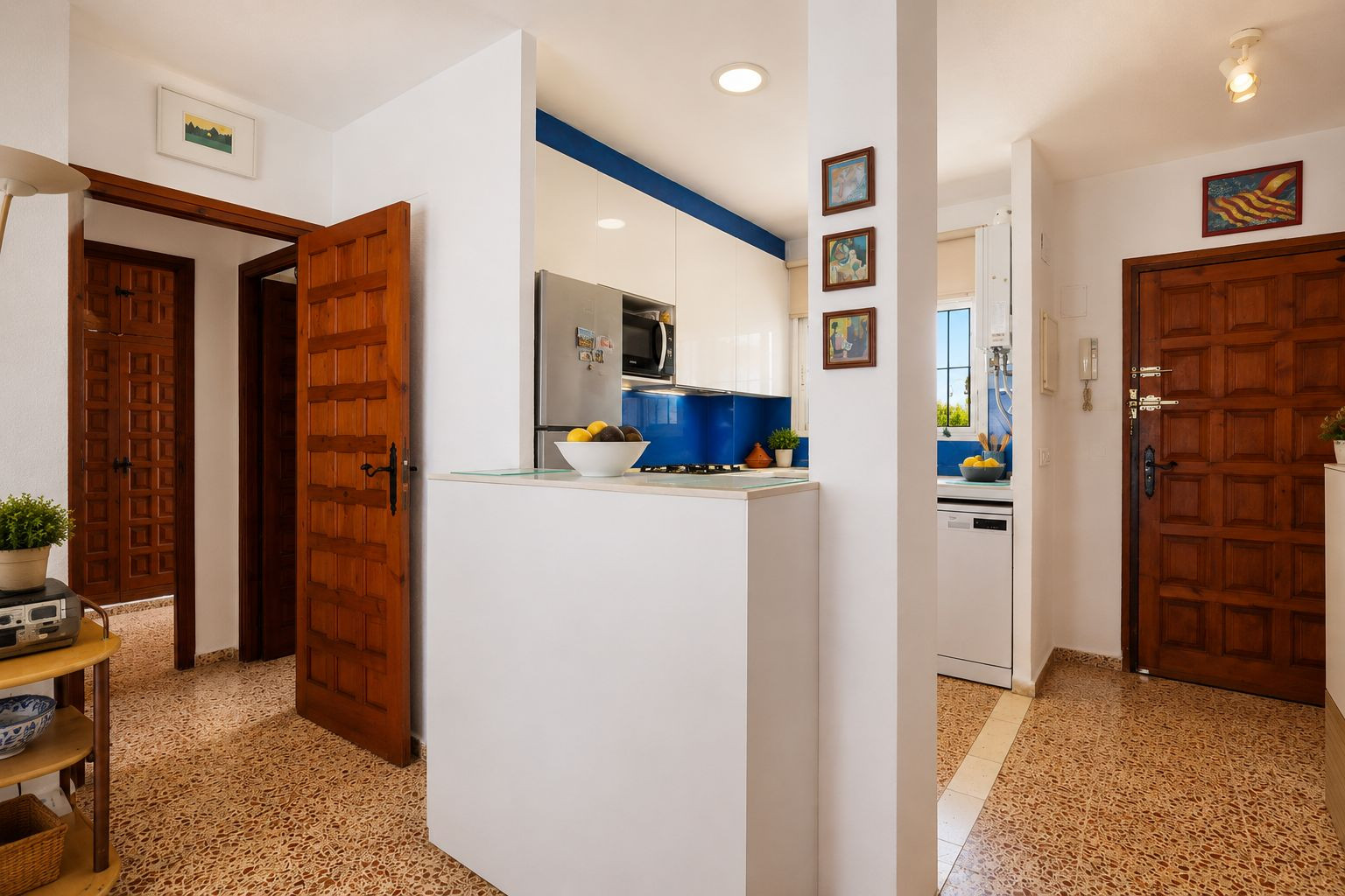 El recibidor presenta un suelo de baldosas con patrón distintivo y puertas de madera clásicas. Una cocina moderna con salpicadero azul y muebles blancos es visible, separada por una práctica barra. El recibidor presenta un suelo de baldosas con patrón distintivo y puertas de madera clásicas. Una cocina moderna con salpicadero azul y muebles blancos es visible, separada por una práctica barra.