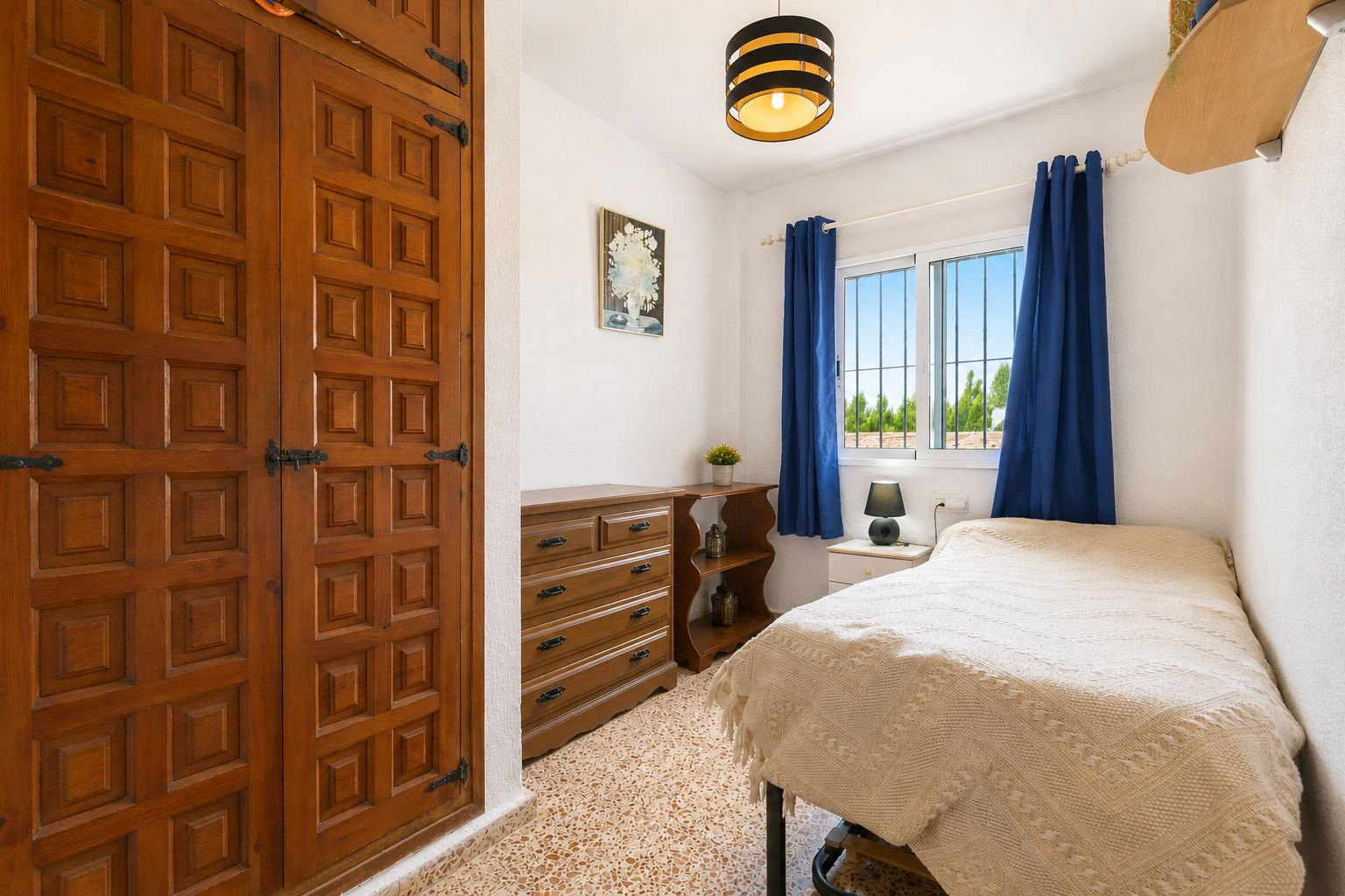 Este acogedor dormitorio presenta un armario empotrado de madera con paneles decorativos y herrajes negros tradicionales. La luz natural entra por una ventana con cortinas azules y vistas a la vegetación. Este acogedor dormitorio presenta un armario empotrado de madera con paneles decorativos y herrajes negros tradicionales. La luz natural entra por una ventana con cortinas azules y vistas a la vegetación.