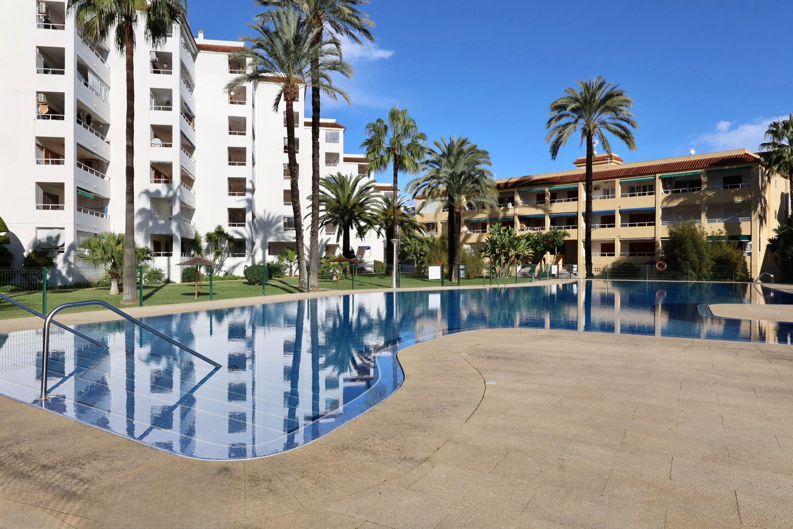La amplia piscina comunitaria con azulejos azules y borde de pavimento claro, incluye barandillas de acero. Palmeras y edificios residenciales se reflejan en sus aguas bajo un cielo azul. La amplia piscina comunitaria con azulejos azules y borde de pavimento claro, incluye barandillas de acero. Palmeras y edificios residenciales se reflejan en sus aguas bajo un cielo azul.