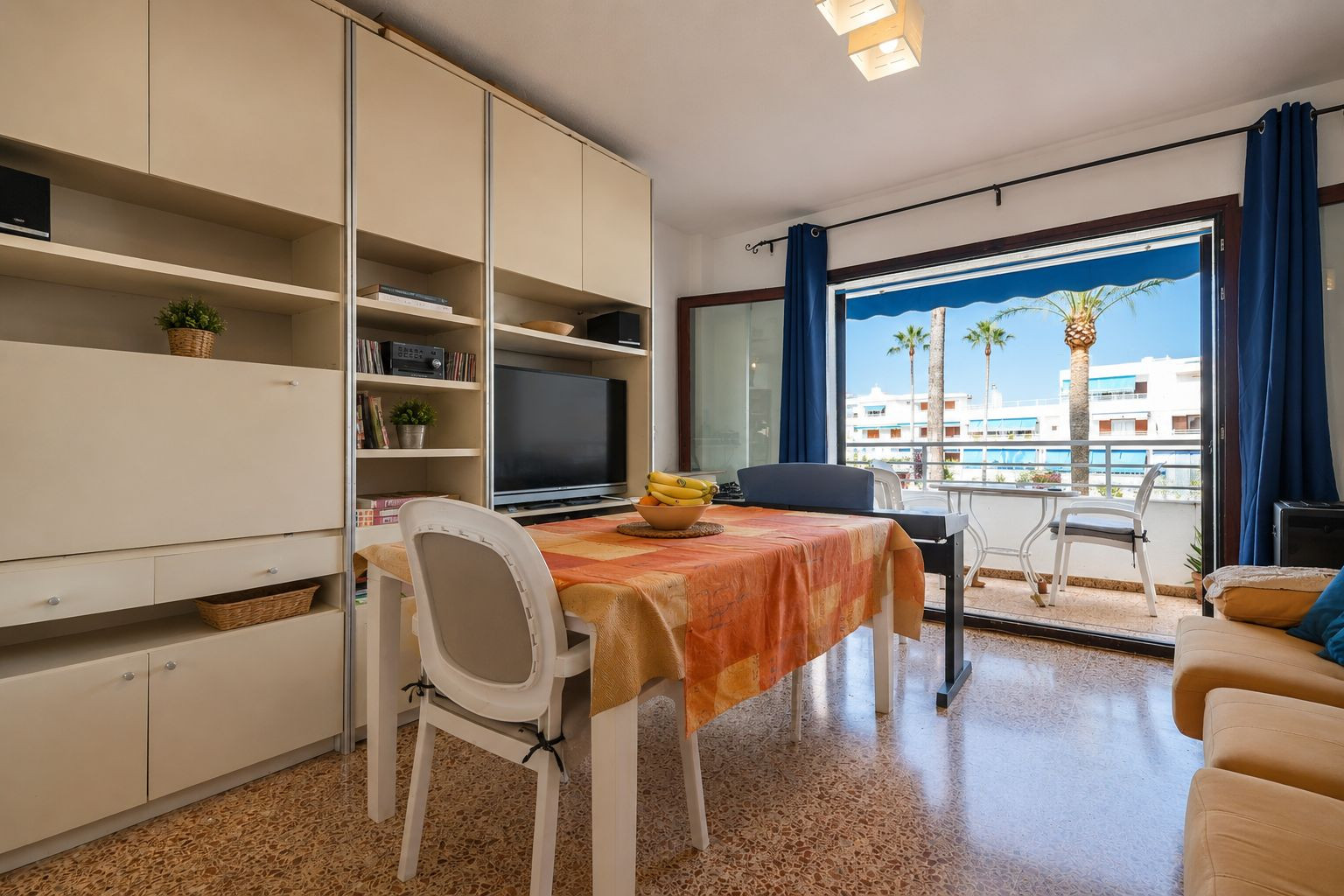 Luminoso salón-comedor con mesa y mueble integrado, que se abre a una terraza exterior con vistas a palmeras y edificios blancos bajo toldos azules. Luminoso salón-comedor con mesa y mueble integrado, que se abre a una terraza exterior con vistas a palmeras y edificios blancos bajo toldos azules.