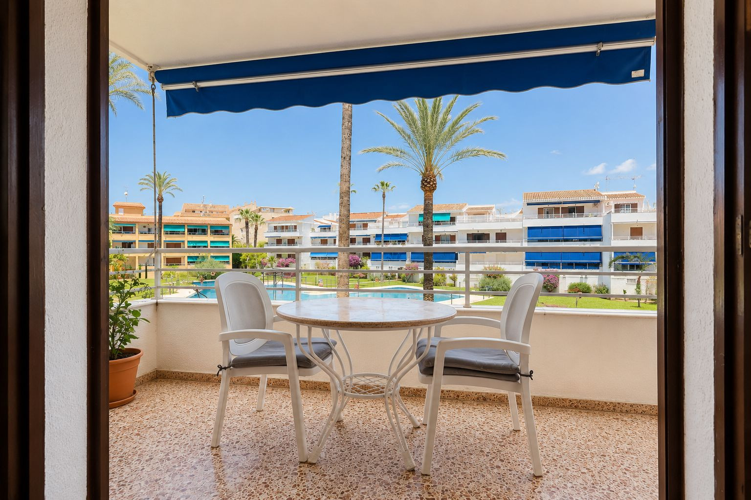 Terraza con suelo de gres, equipada con mesa y dos sillas, bajo un toldo azul. Ofrece vistas directas a la piscina comunitaria, jardines y palmeras. Terraza con suelo de gres, equipada con mesa y dos sillas, bajo un toldo azul. Ofrece vistas directas a la piscina comunitaria, jardines y palmeras.