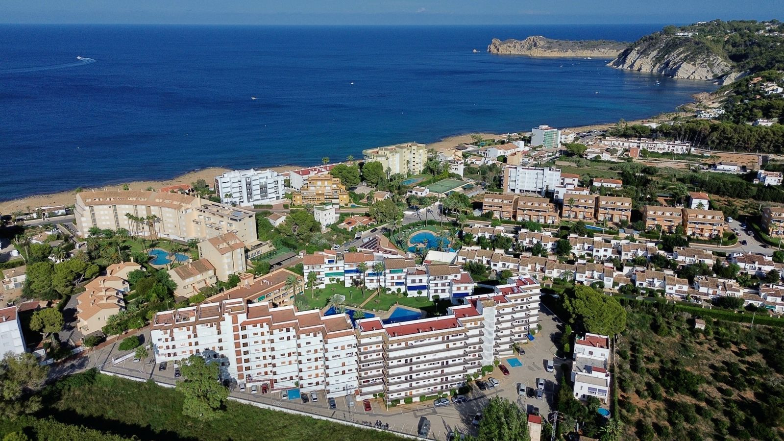 Vista aérea de una urbanización costera con múltiples edificios residenciales, piscinas comunitarias y zonas verdes, junto a una playa de arena y el Mediterráneo, con un cabo rocoso al fondo. Vista aérea de una urbanización costera con múltiples edificios residenciales, piscinas comunitarias y zonas verdes, junto a una playa de arena y el Mediterráneo, con un cabo rocoso al fondo.