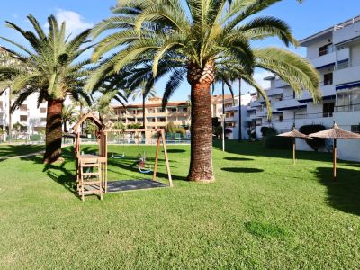 Amplio jardín comunitario con césped cuidado, parque infantil de madera, palmeras y sombrillas de paja, rodeado de edificios residenciales.