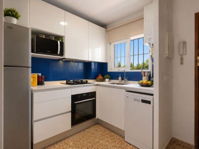 Cocina moderna y funcional con armarios blancos brillantes, encimera clara y salpicadero azul. Equipada con electrodomésticos integrados y ventana con vistas a la vegetación.