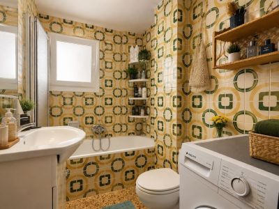 El baño completo presenta azulejos con un distintivo patrón geométrico en tonos amarillo y verde, un lavabo con mueble, bañera con mampara y lavadora integrada, iluminado por una ventana esmerilada.