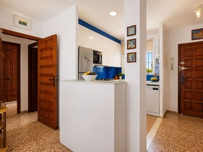 El recibidor presenta un suelo de baldosas con patrón distintivo y puertas de madera clásicas. Una cocina moderna con salpicadero azul y muebles blancos es visible, separada por una práctica barra.
