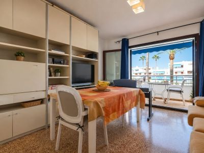 Luminoso salón-comedor con mesa y mueble integrado, que se abre a una terraza exterior con vistas a palmeras y edificios blancos bajo toldos azules.