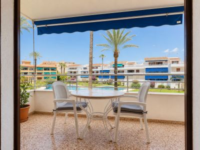 Terraza con suelo de gres, equipada con mesa y dos sillas, bajo un toldo azul. Ofrece vistas directas a la piscina comunitaria, jardines y palmeras.