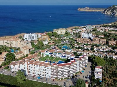 Vista aérea de una urbanización costera con múltiples edificios residenciales, piscinas comunitarias y zonas verdes, junto a una playa de arena y el Mediterráneo, con un cabo rocoso al fondo.