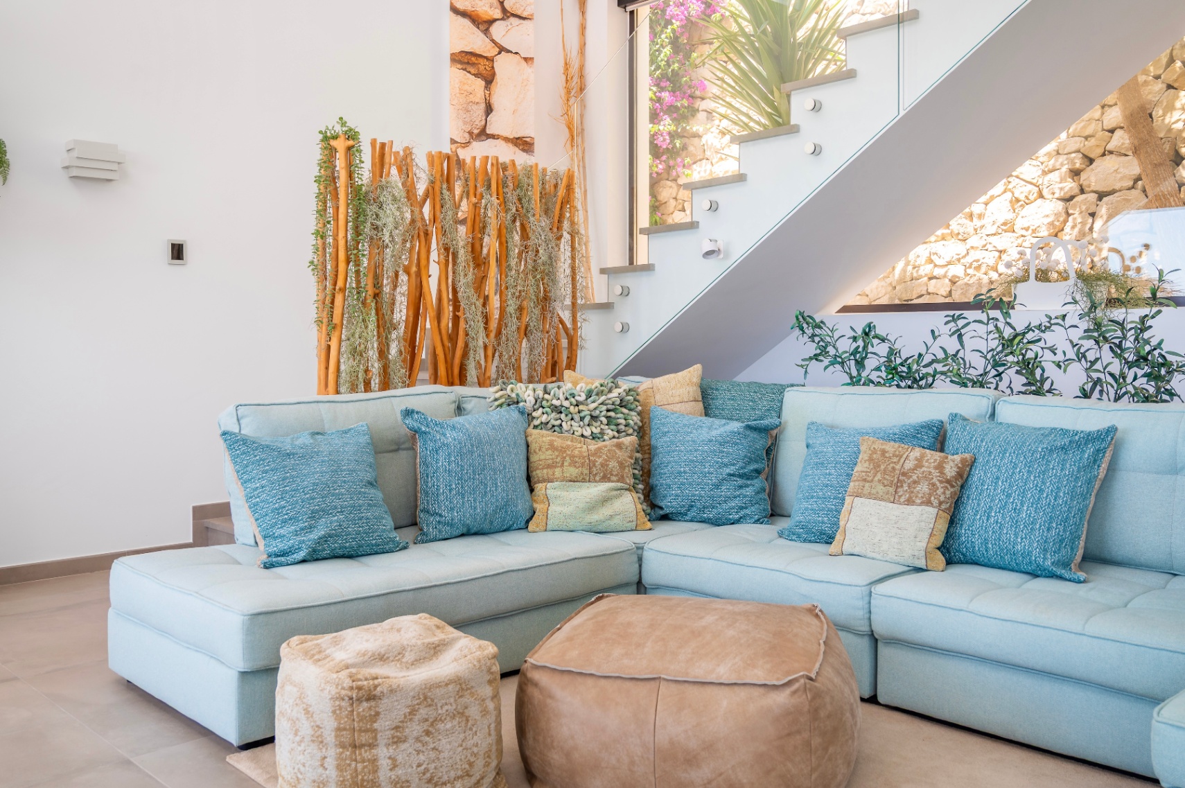 Acogedora sala de estar con sofá seccional azul claro, cojines variados y dos pufs de cuero. Escalera moderna de cristal junto a una pantalla de madera y plantas, con muro de piedra visible. Acogedora sala de estar con sofá seccional azul claro, cojines variados y dos pufs de cuero. Escalera moderna de cristal junto a una pantalla de madera y plantas, con muro de piedra visible.
