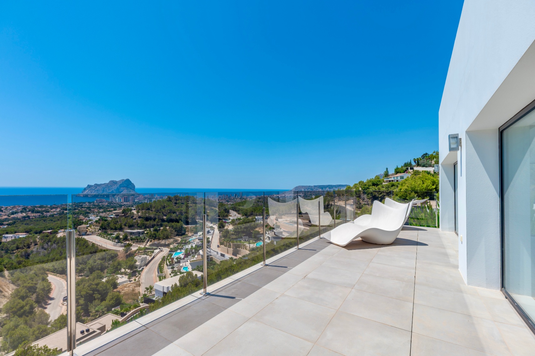 Amplia terraza exterior con suelo de baldosas claras, una moderna tumbona blanca y barandilla de cristal. Vistas panorámicas al mar Mediterráneo, el Peñón de Ifach y la costa de Calpe. Amplia terraza exterior con suelo de baldosas claras, una moderna tumbona blanca y barandilla de cristal. Vistas panorámicas al mar Mediterráneo, el Peñón de Ifach y la costa de Calpe.