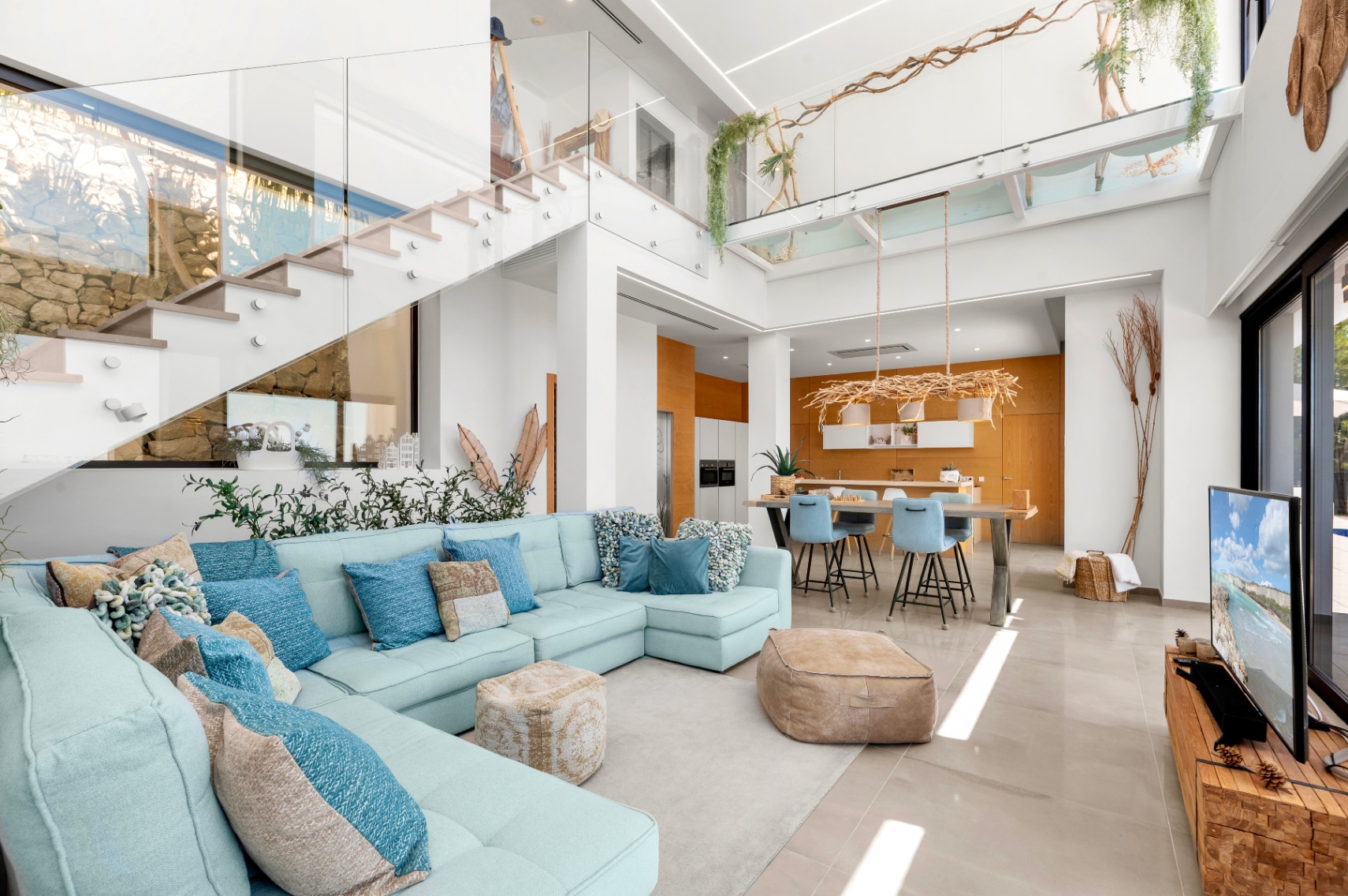 Amplio salón de planta abierta con sofá seccional azul claro, escalera moderna de cristal y cocina con detalles de madera cálida, todo iluminado por luz natural. Amplio salón de planta abierta con sofá seccional azul claro, escalera moderna de cristal y cocina con detalles de madera cálida, todo iluminado por luz natural.