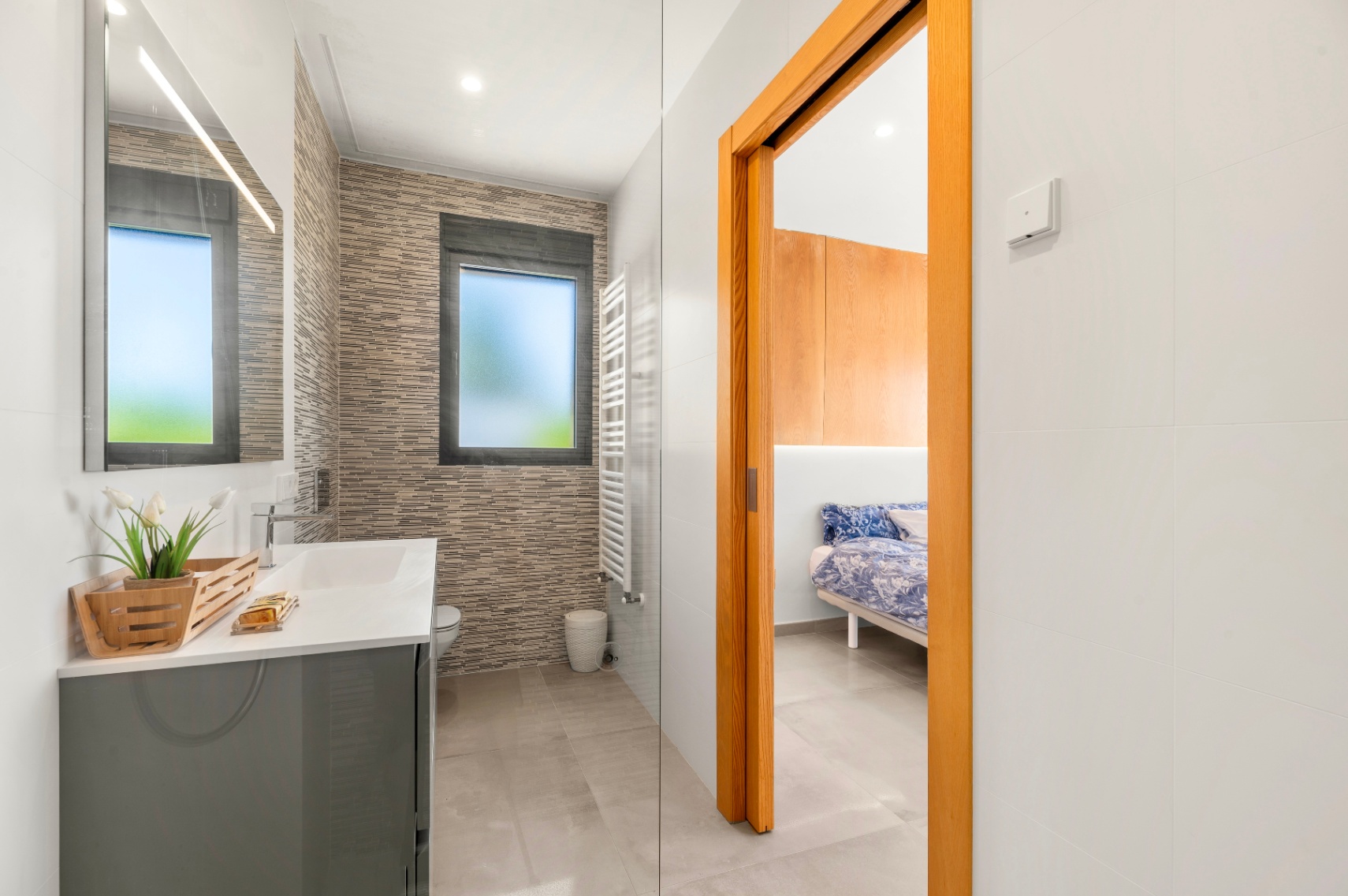 Baño moderno con lavabo gris, espejo iluminado y azulejos texturizados. Incluye una ventana esmerilada, toallero calefactado y acceso directo a un dormitorio. Baño moderno con lavabo gris, espejo iluminado y azulejos texturizados. Incluye una ventana esmerilada, toallero calefactado y acceso directo a un dormitorio.