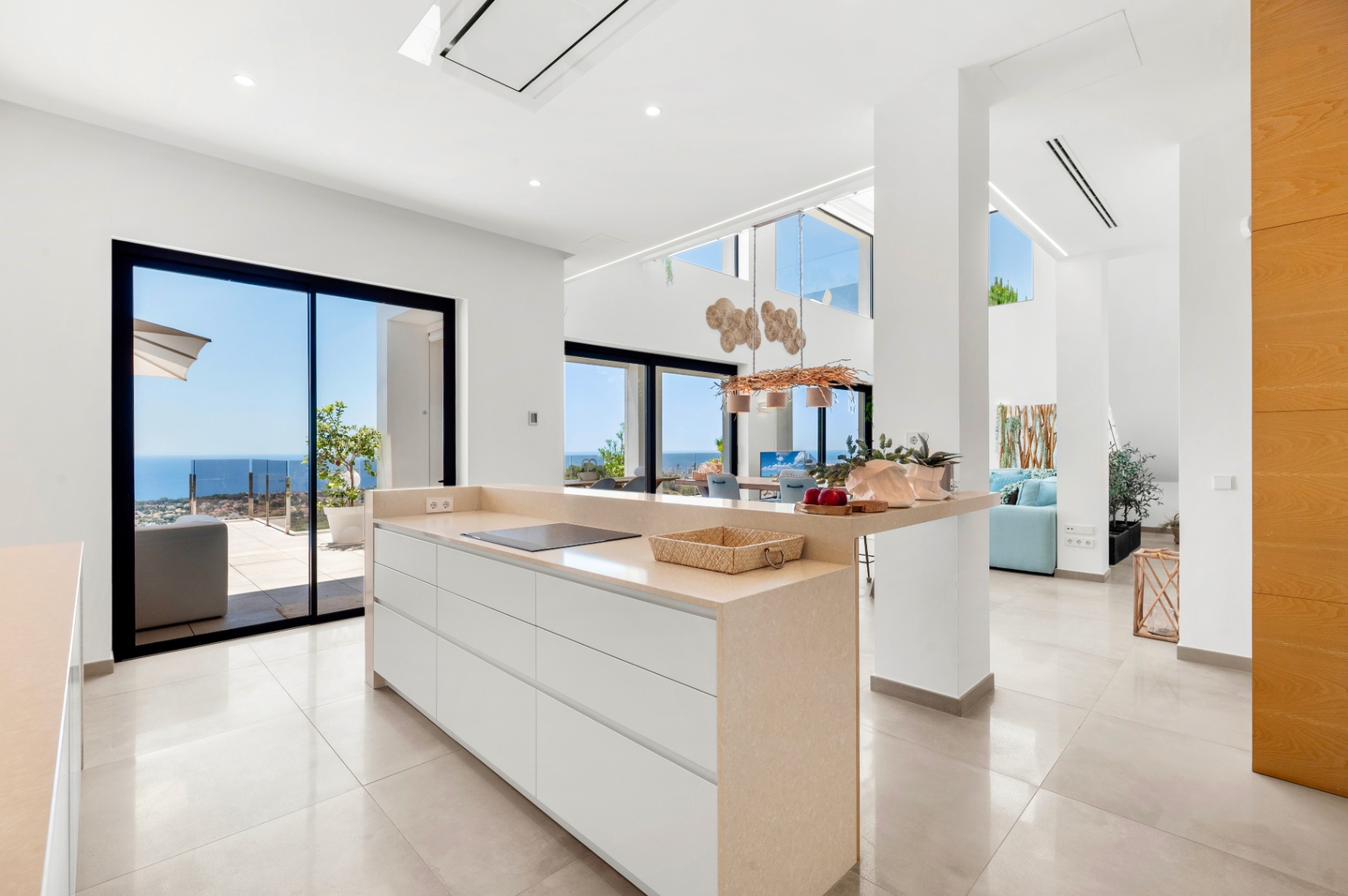 Cocina de diseño abierta con isla central de encimera beige y placa de inducción. Grandes ventanales correderos ofrecen vistas directas al mar Mediterráneo desde un espacio luminoso y moderno. Cocina de diseño abierta con isla central de encimera beige y placa de inducción. Grandes ventanales correderos ofrecen vistas directas al mar Mediterráneo desde un espacio luminoso y moderno.