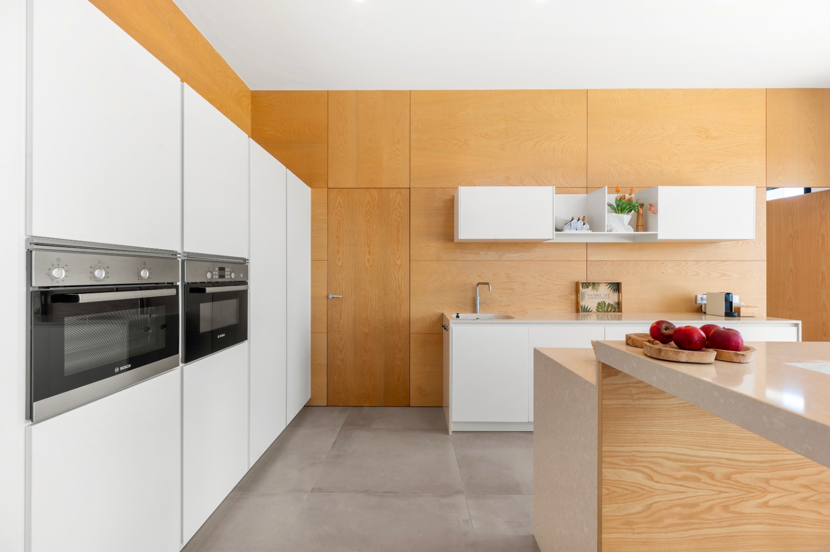Cocina moderna con armarios blancos integrados y hornos Bosch. Destaca la isla central con encimera de piedra clara y el revestimiento de madera clara en paredes y techo. Cocina moderna con armarios blancos integrados y hornos Bosch. Destaca la isla central con encimera de piedra clara y el revestimiento de madera clara en paredes y techo.