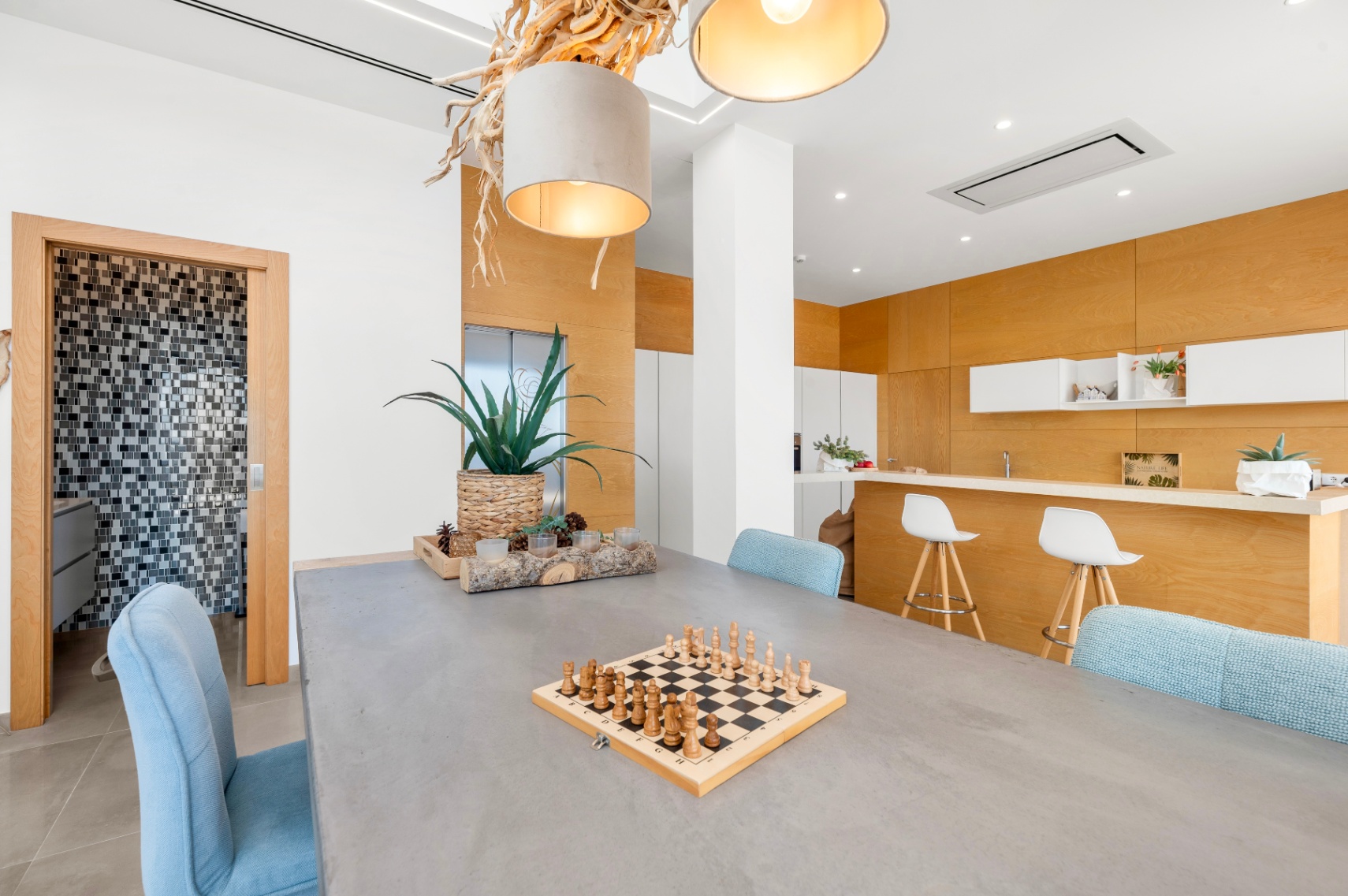 Comedor moderno con mesa de hormigón y sillas azules. Destaca una lámpara colgante de madera natural y una cocina abierta con panelado de madera clara y barra. Al fondo, un baño con mosaicos. Comedor moderno con mesa de hormigón y sillas azules. Destaca una lámpara colgante de madera natural y una cocina abierta con panelado de madera clara y barra. Al fondo, un baño con mosaicos.