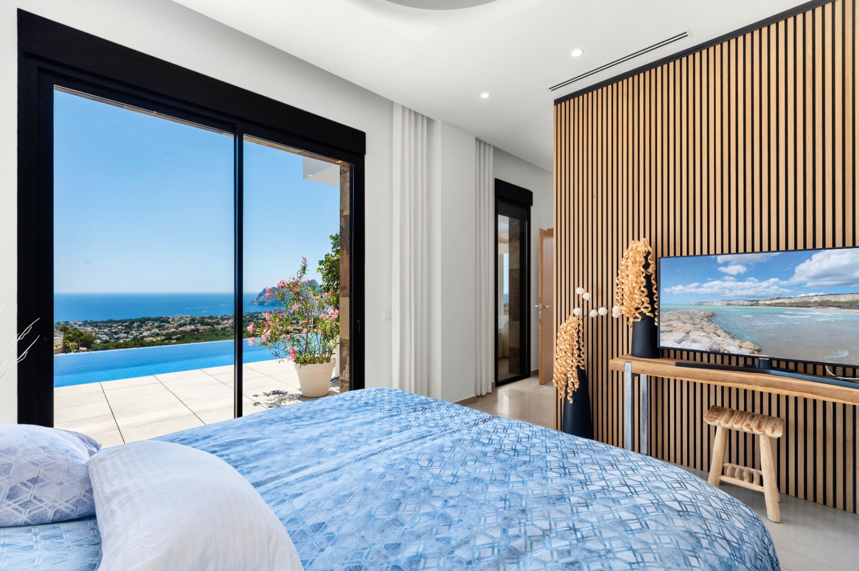 Dormitorio con amplias puertas correderas de cristal que se abren a una piscina infinita y vistas panorámicas al mar. Una moderna pared de lamas de madera enmarca la televisión, complementando el interior luminoso. Dormitorio con amplias puertas correderas de cristal que se abren a una piscina infinita y vistas panorámicas al mar. Una moderna pared de lamas de madera enmarca la televisión, complementando el interior luminoso.