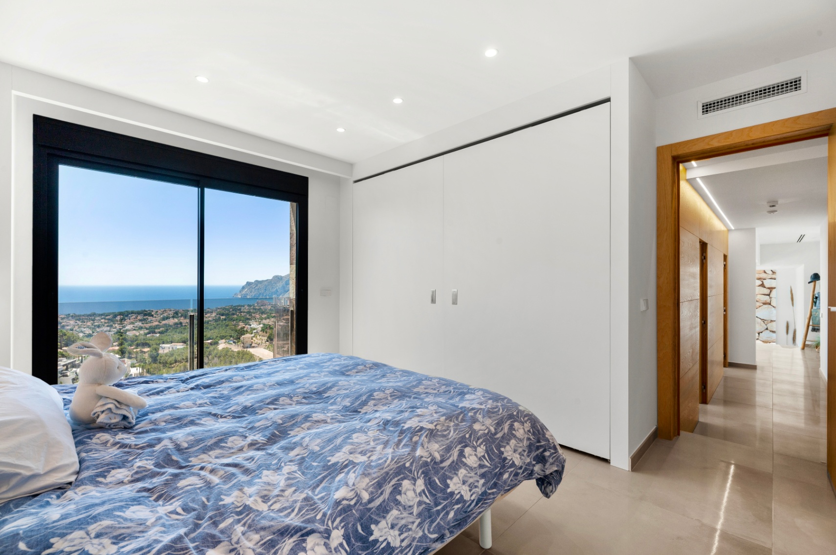 Dormitorio con puertas correderas de cristal de suelo a techo ofreciendo vistas panorámicas al mar y la ciudad. Incluye armarios empotrados blancos y suelo porcelánico claro. Dormitorio con puertas correderas de cristal de suelo a techo ofreciendo vistas panorámicas al mar y la ciudad. Incluye armarios empotrados blancos y suelo porcelánico claro.