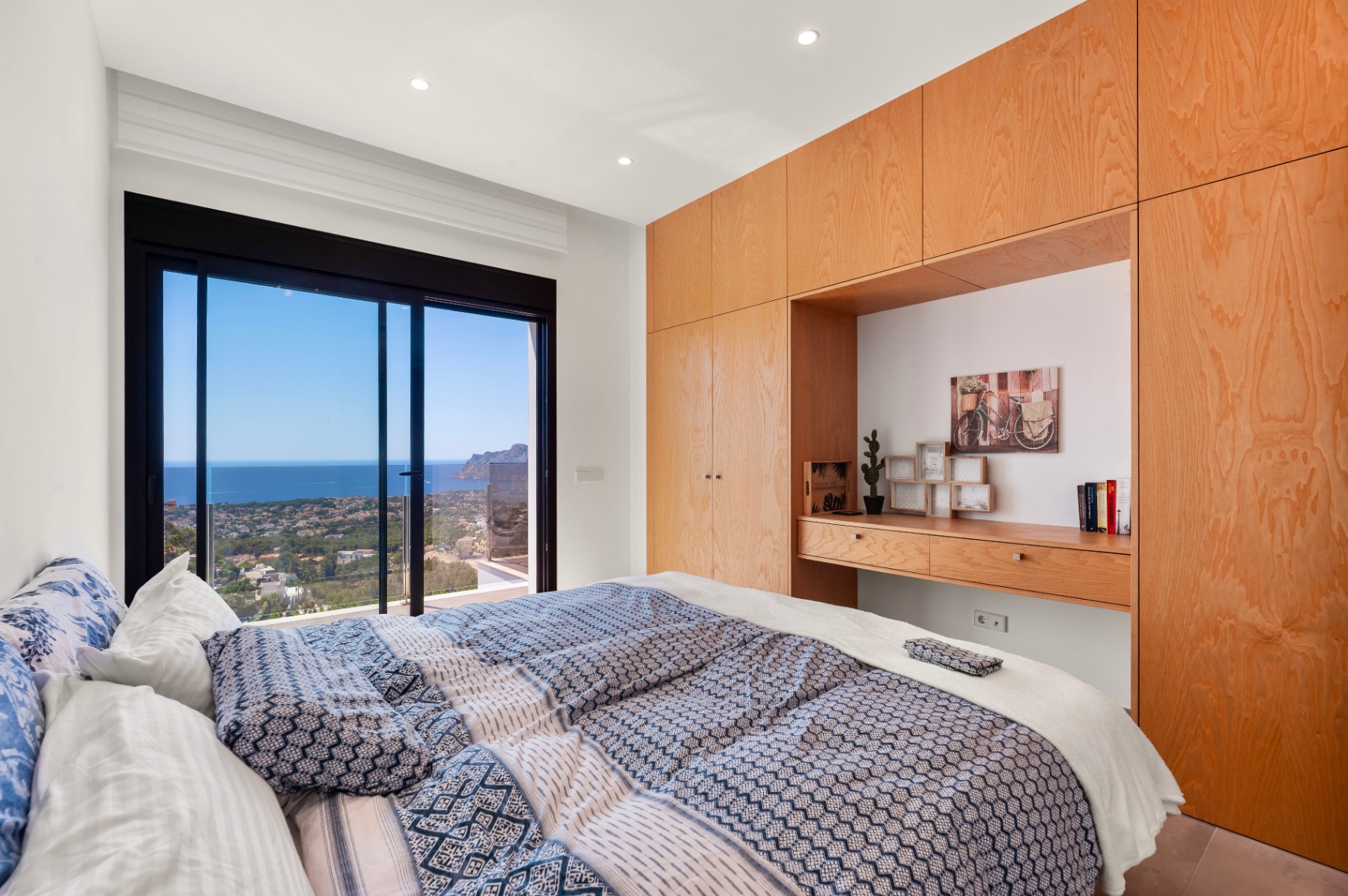 Dormitorio principal con amplias puertas correderas de cristal y vistas panorámicas al mar y la costa. Armarios empotrados de madera clara con escritorio integrado. Dormitorio principal con amplias puertas correderas de cristal y vistas panorámicas al mar y la costa. Armarios empotrados de madera clara con escritorio integrado.