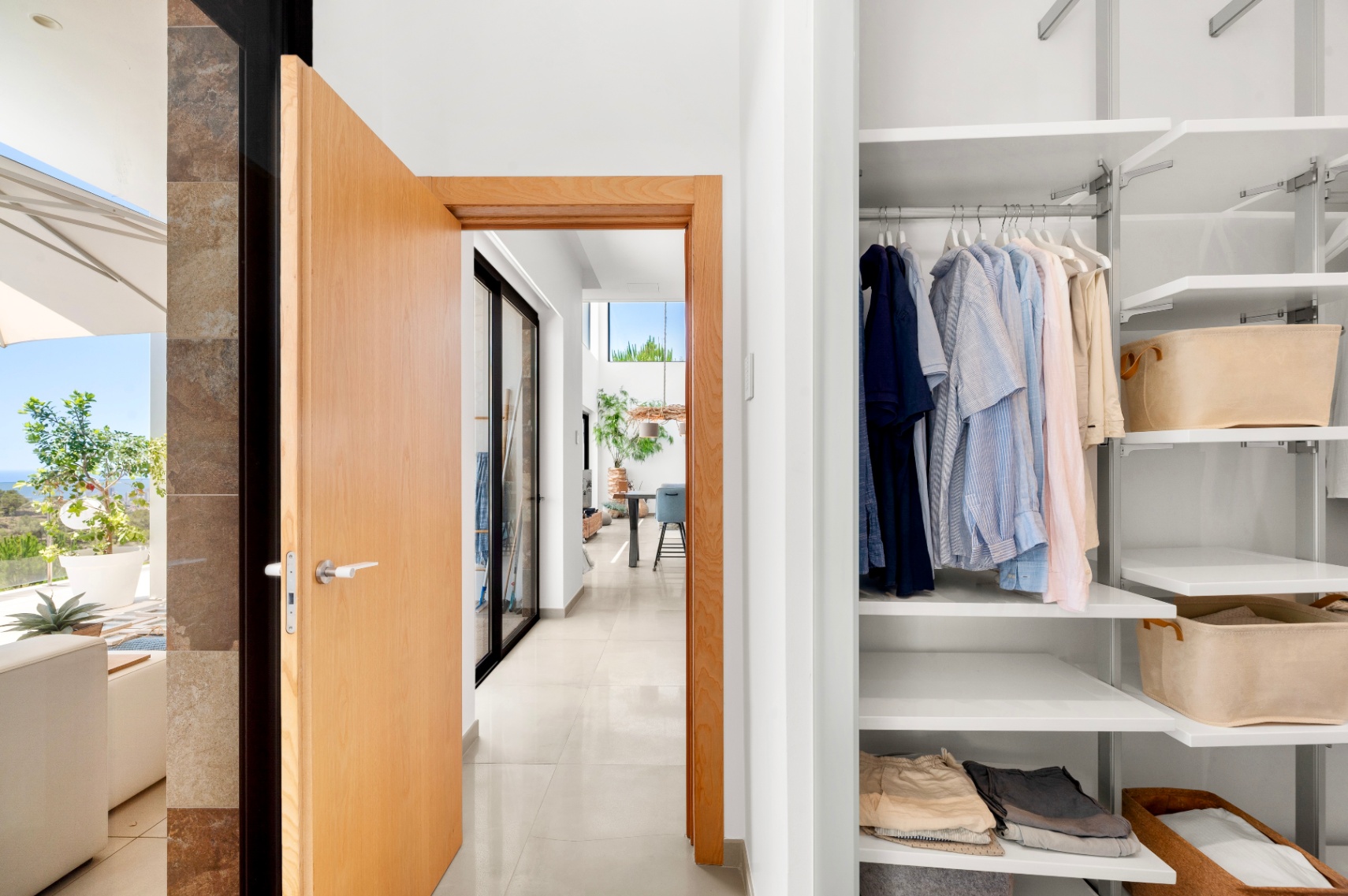 Entrada a un moderno pasillo con puerta de madera clara abierta, mostrando un armario empotrado blanco con estantes ajustables y una terraza exterior soleada con vistas al mar. Entrada a un moderno pasillo con puerta de madera clara abierta, mostrando un armario empotrado blanco con estantes ajustables y una terraza exterior soleada con vistas al mar.