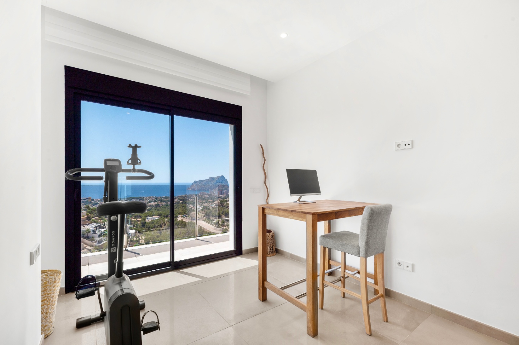 Espacio multifuncional con escritorio alto de madera y bicicleta estática, ambos orientados a las amplias vistas al mar y al Peñon de Ifach a través de las grandes puertas correderas de cristal. Espacio multifuncional con escritorio alto de madera y bicicleta estática, ambos orientados a las amplias vistas al mar y al Peñon de Ifach a través de las grandes puertas correderas de cristal.
